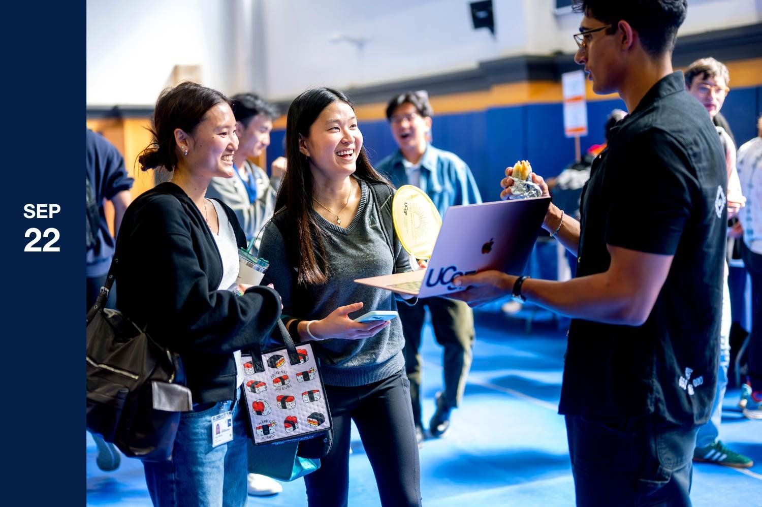 The UCSF mascot, a brown bear, poses enthusiastically next to two smiling students at a crowded indoor event. The student in the center is laughing as she looks at the student next to her.