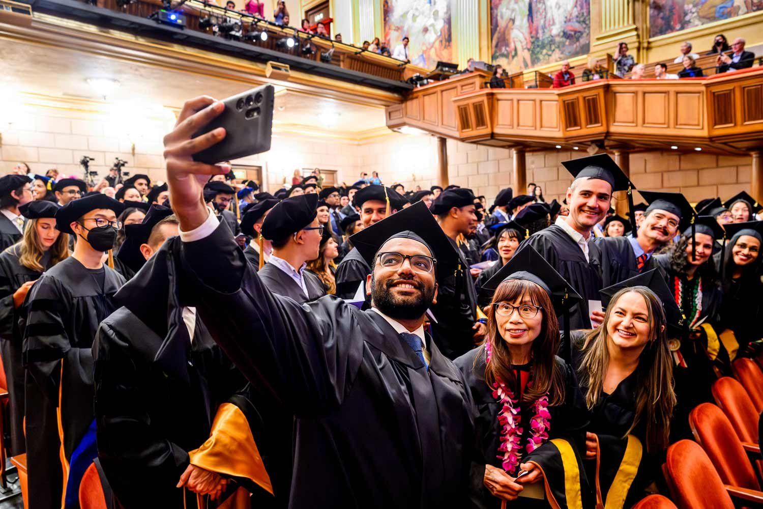 Graduates at a commencement ceremony smile as they pose for a selfie.
