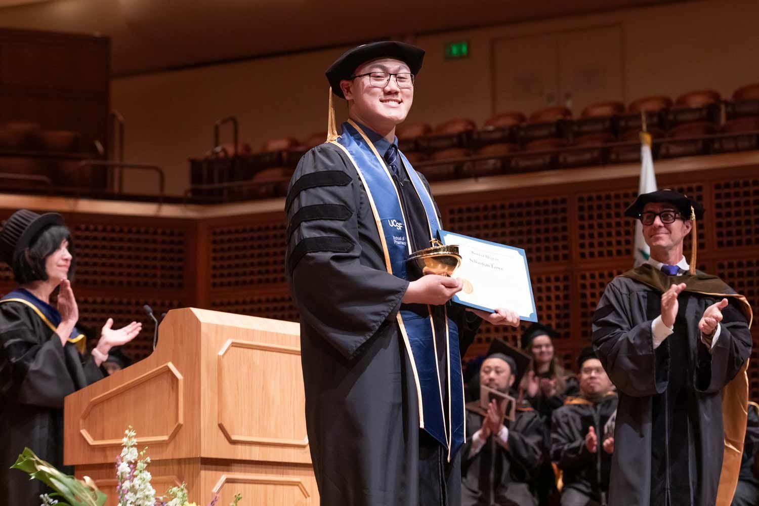 A graduate in regalia holds an award onstage.