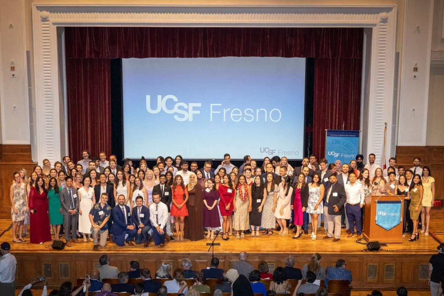 Residents and fellows standing together on stage under bright auditorium lighting during a commencement ceremony.