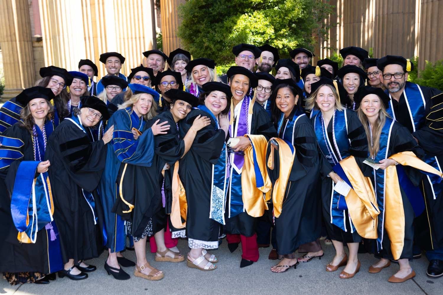 A large group of UCSF School of Nursing graduates in blue academic regalia and caps smile and celebrate together in an outdoor crowd.