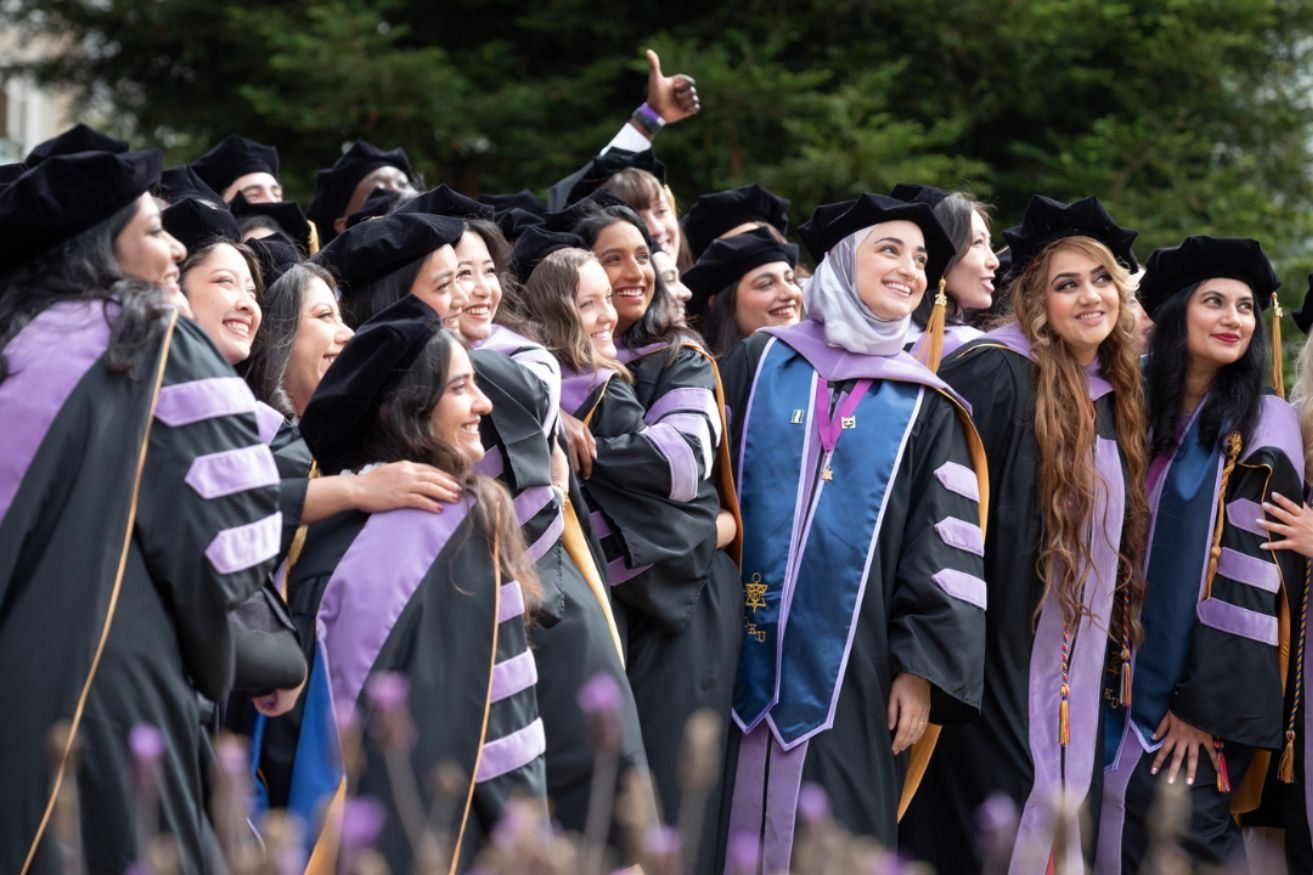 Large group of graduates, mostly women, smile in robes outdoors.