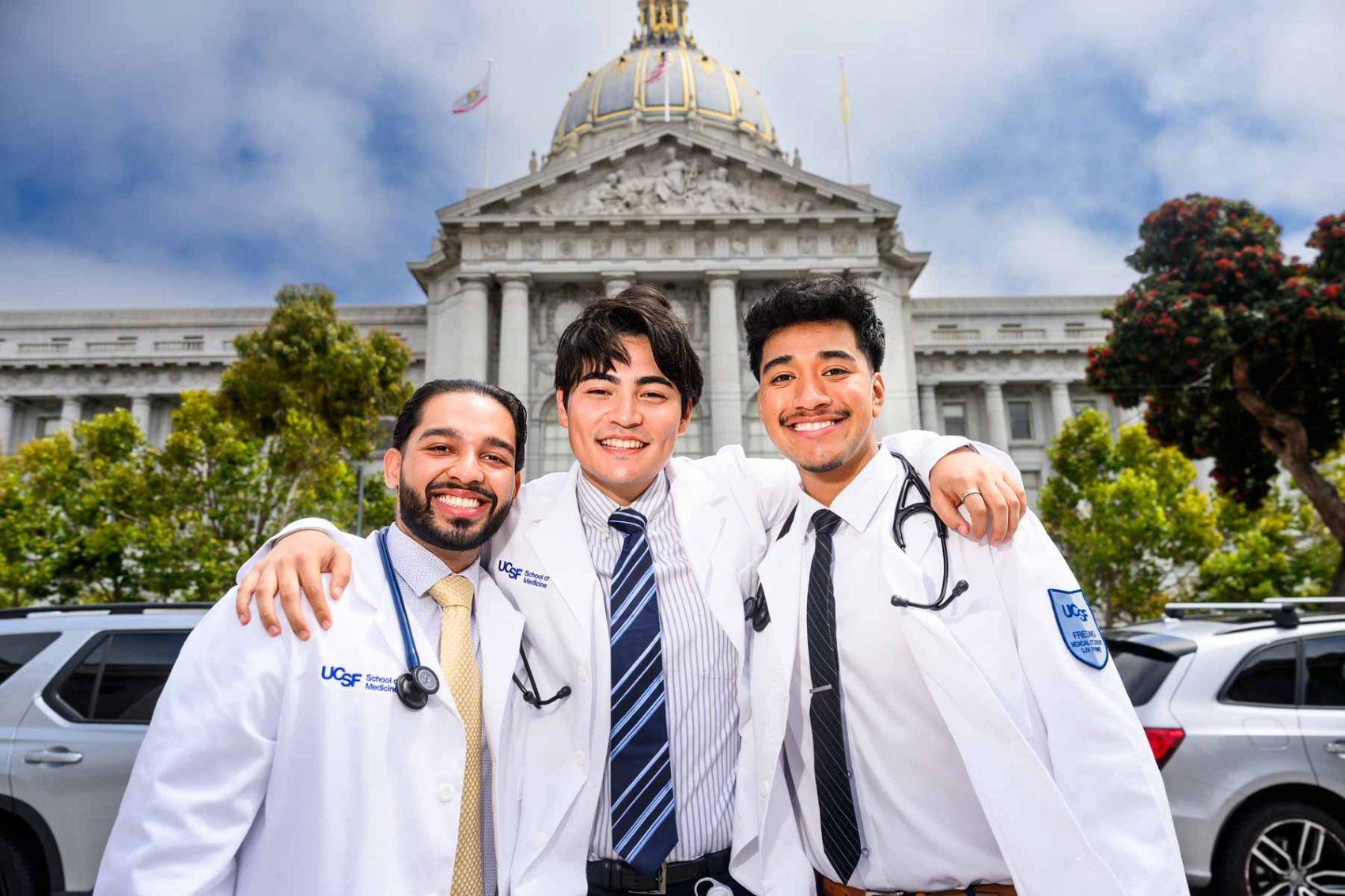 Three medical students wearing white coats and stethoscopes pose together outdoors.