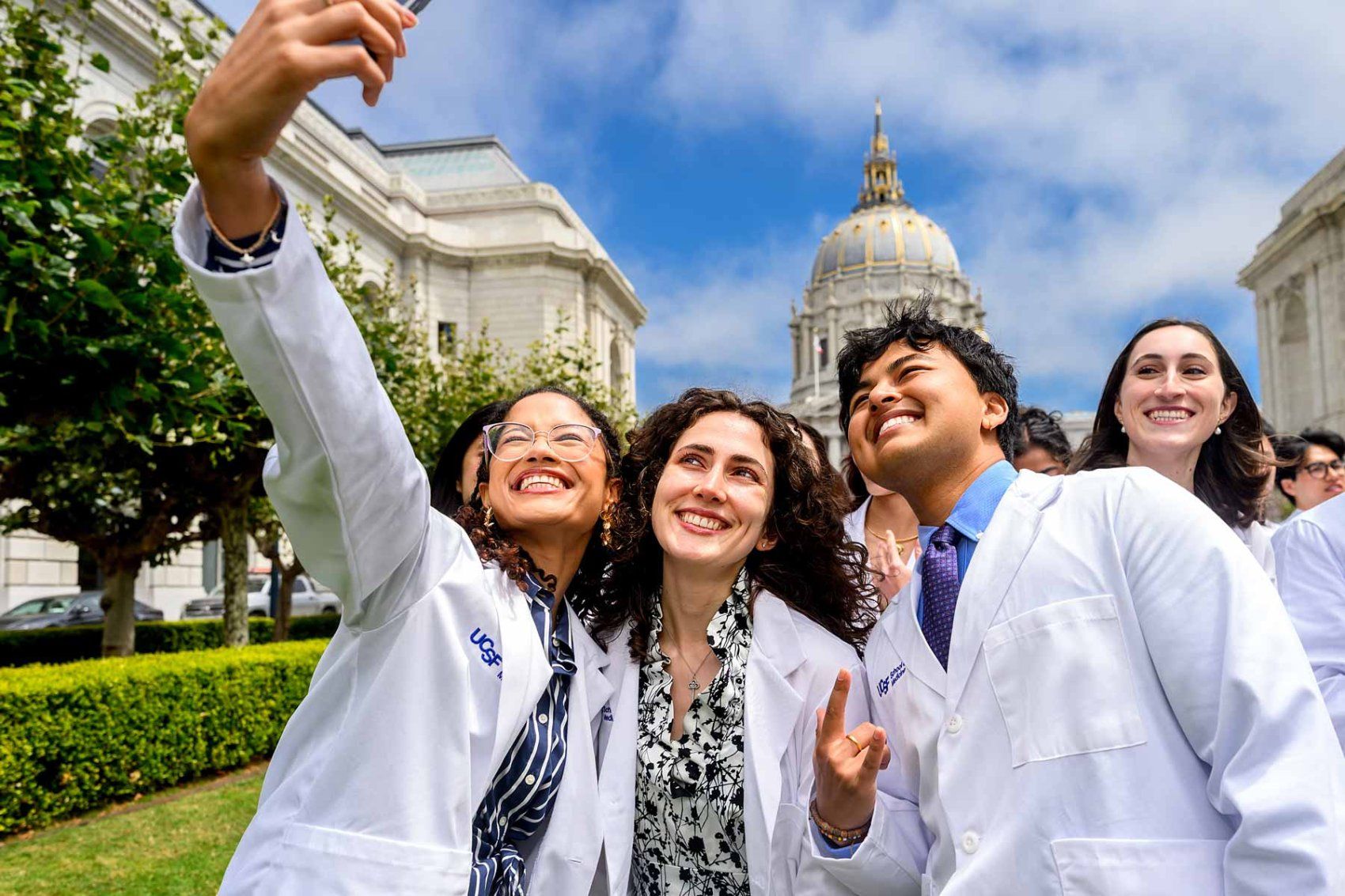 Four School of Medicine students take a selfie outdoors wearing white coats.