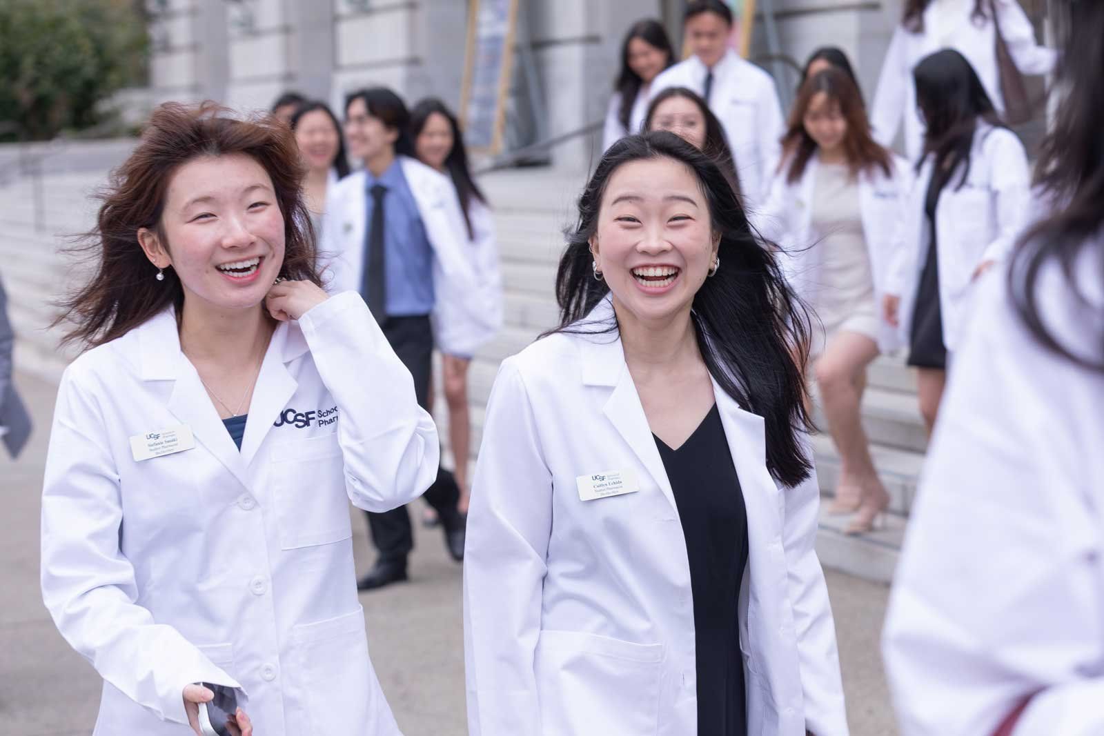 UCSF School of Pharmacy students in white coats smile and walk together outdoors after a ceremony.