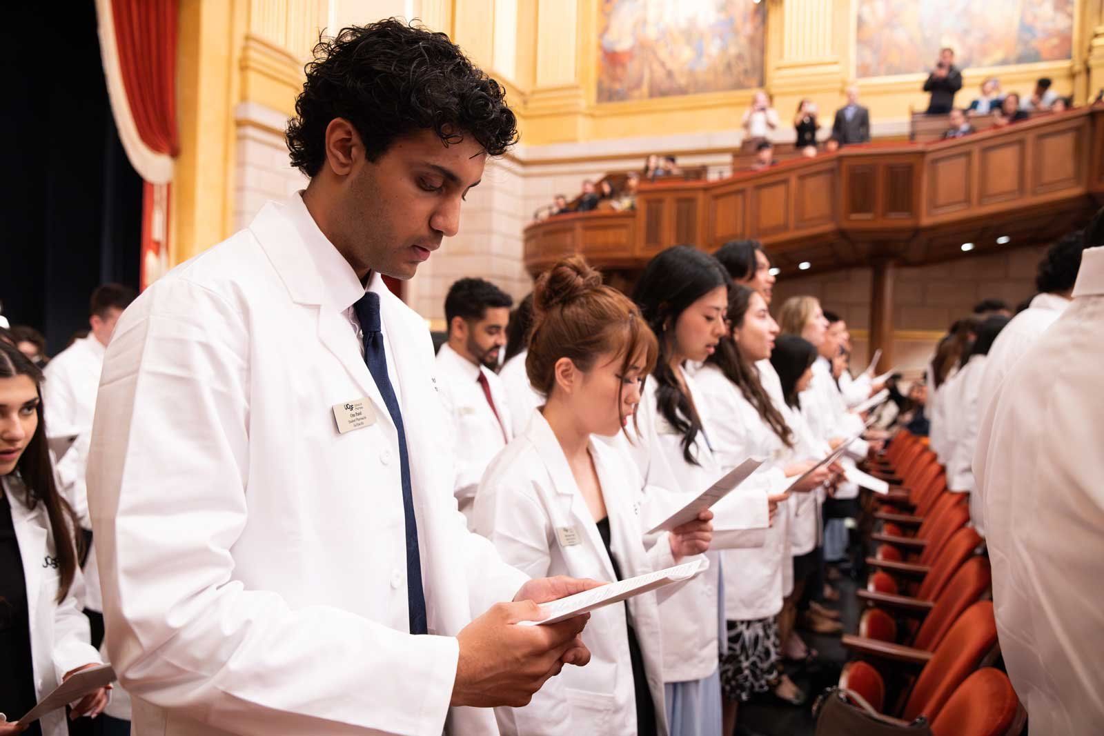 U C S F School of Pharmacy students wearing white coats stand together and read the pharmacist’s oath during a ceremony.