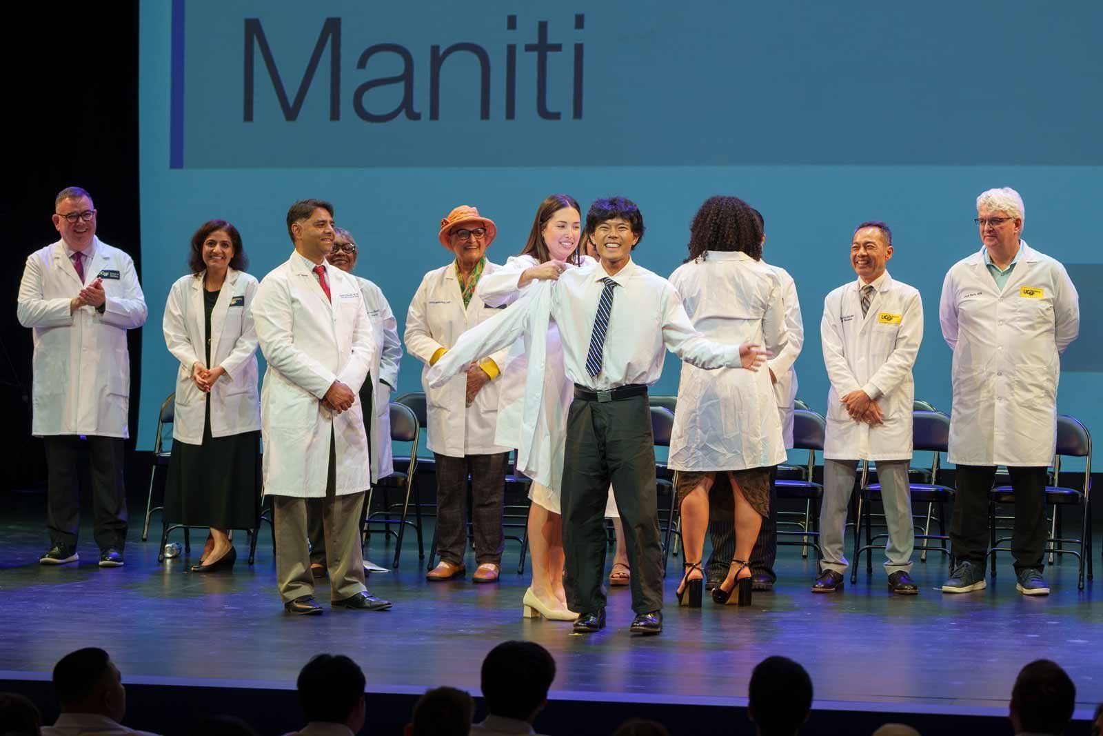 A dental student receives his white coat on stage as faculty on stage watch.