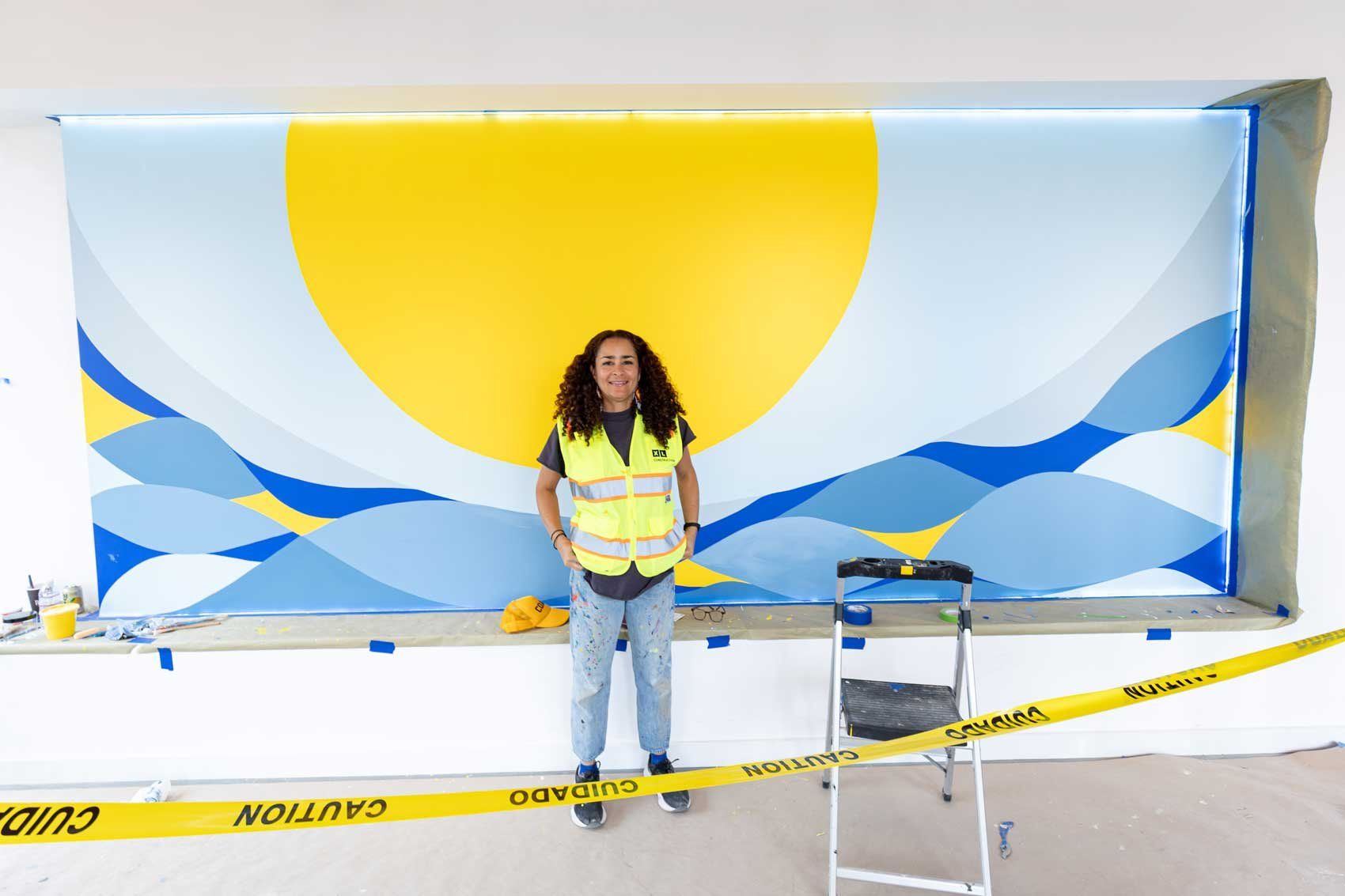 Artist Adia Millett stands in front of a painted mural showing a large yellow circle and waves in different shades of blue.