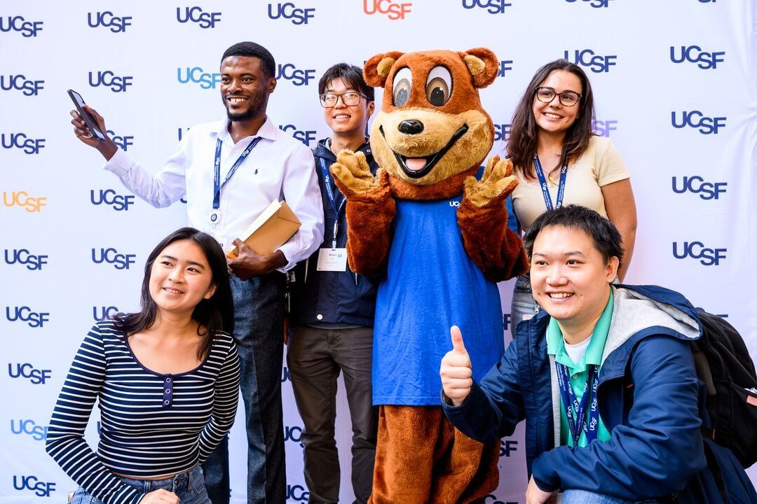 A diverse group of graduate students pose with the U C S F bear mascot in front of a backdrop with the UCSF logo.