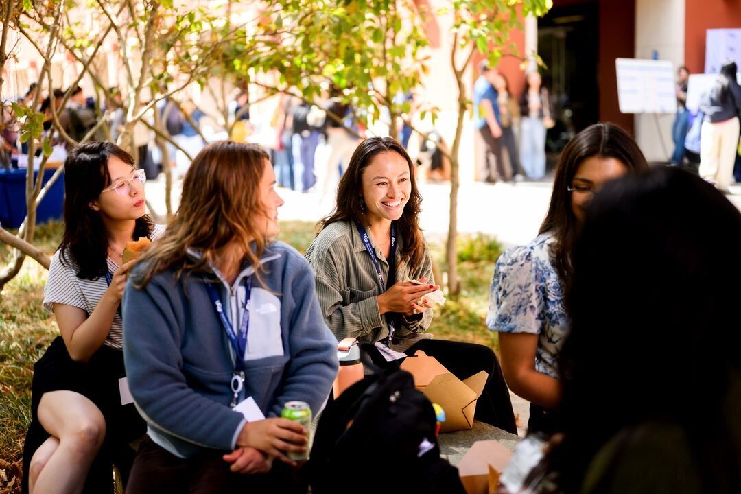 Five female graduate students smile as they sit together and eat outside.