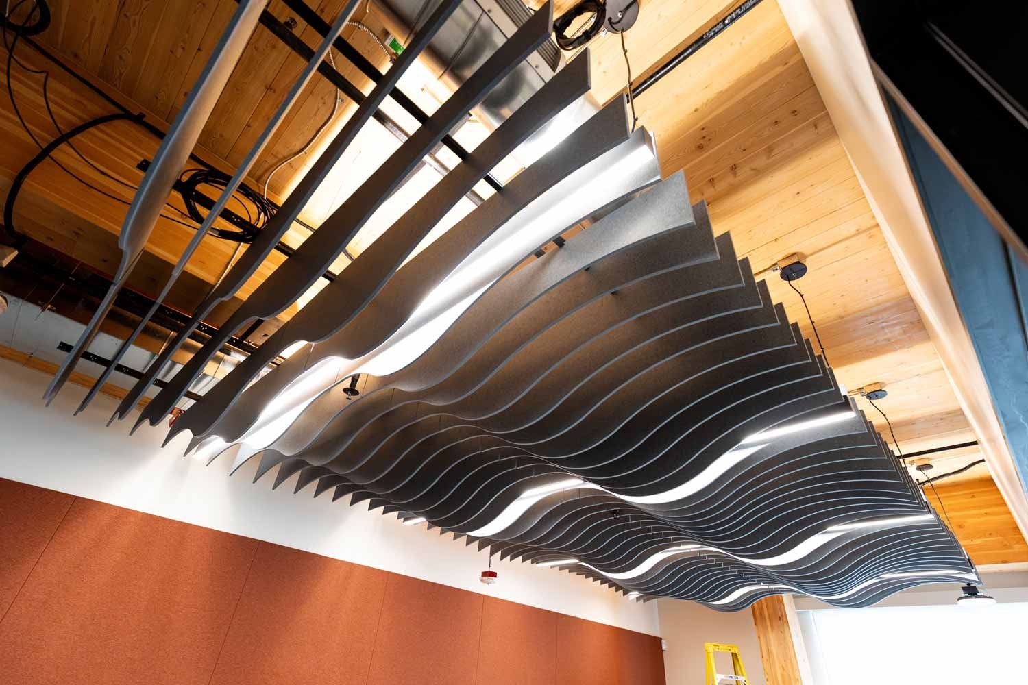 Looking upward at a sculptural installation of suspended, wave-shaped panels beneath the exposed wood ceiling of Temescal Hall.