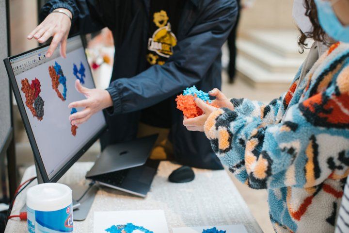 A young attendee holds blue and red 3D models of proteins, while an instructor shows her how they connect on a computer screen