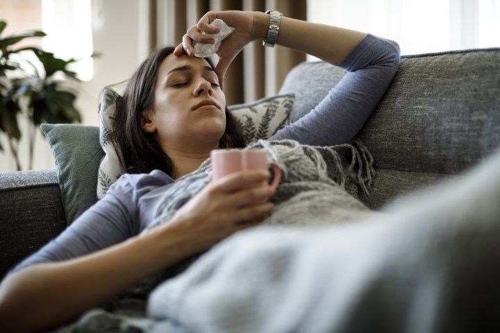 Sick woman lying on her couch, holding a mug and a tissue