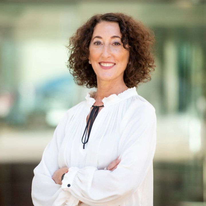 Judith Auerbach stands with her arms crossed in front of glass doors. She wears a white shirt