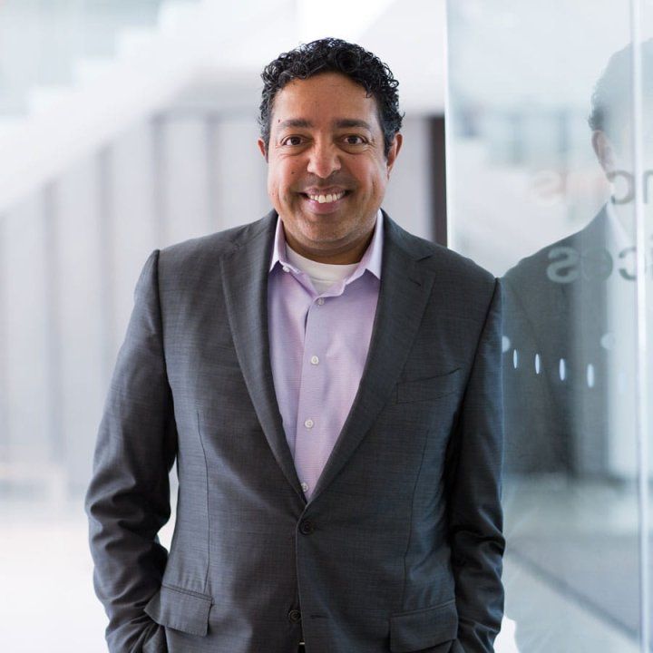 Atul Butte poses for a portrait in a light-filled room with white stairs in the background. He leans against a glass panel