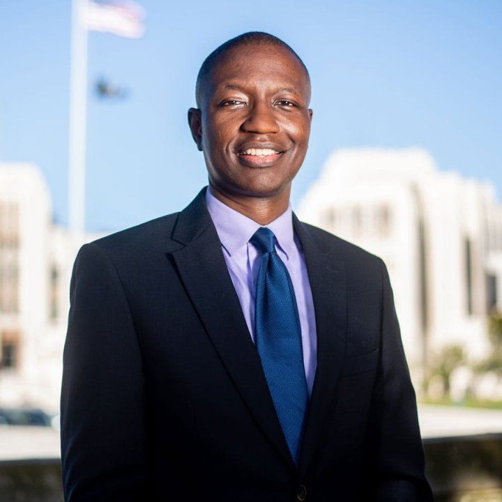 Bruce Ovbiagele, wearing a navy stuit and blue tie, smiles as he poses for a portrait in front of the San Francisco VA Medical Center. An American flag waves on a flagpole.