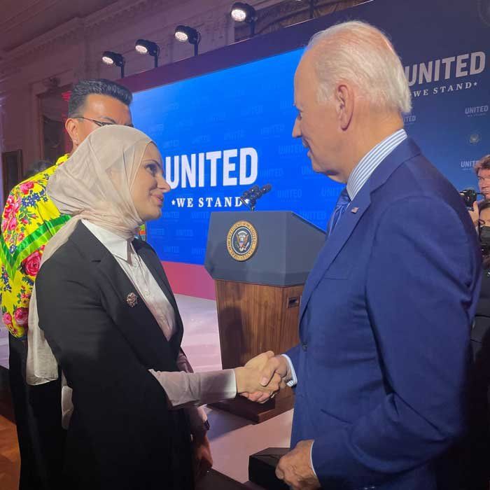 Suzanne Barakat (left) shakes hands with President Joe Biden (right). In the background is a podium and a screen that reads "United We Stand."