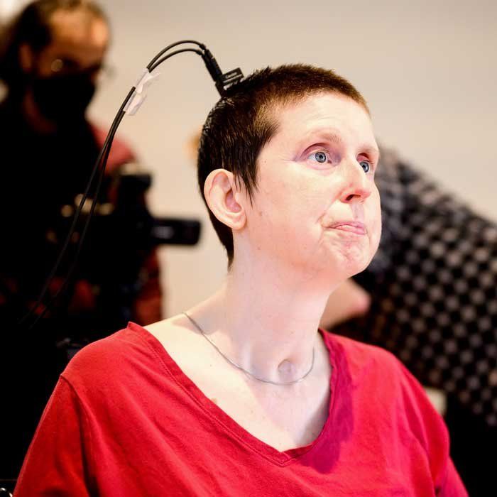 A woman wearing a red shirt and with a buzz haircut sits calmly as an implanted device on her skull is connected to wires.
