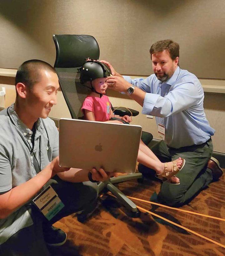 Kevin Bender (right) kneels as he adjusts a helmet on a young girl's head. She is sitting on an office chair, and the helmet will measure her eye movement. Chenyu Want (left) kneels and holds a laptop.