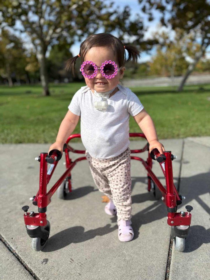 A toddler girl named Carson smiles as she stands with a small walker and a trachial tube.