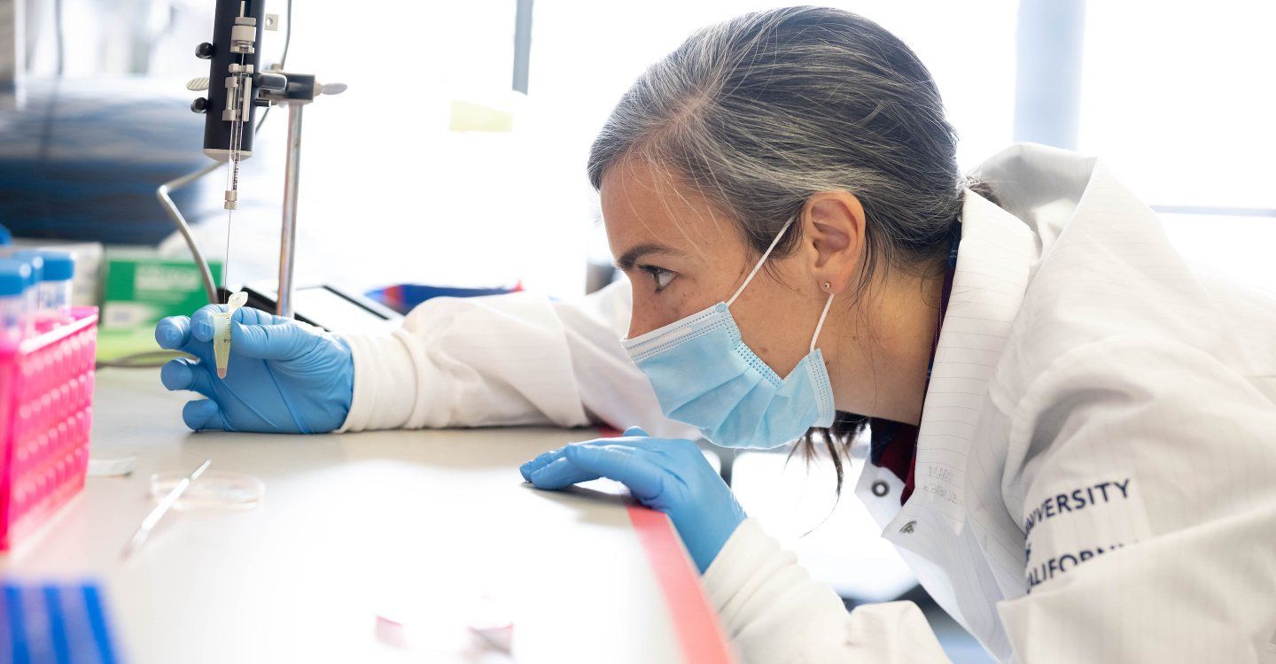 Researcher wearing gloves leans her chin against a lab bench while she looks at a test tube she's holding