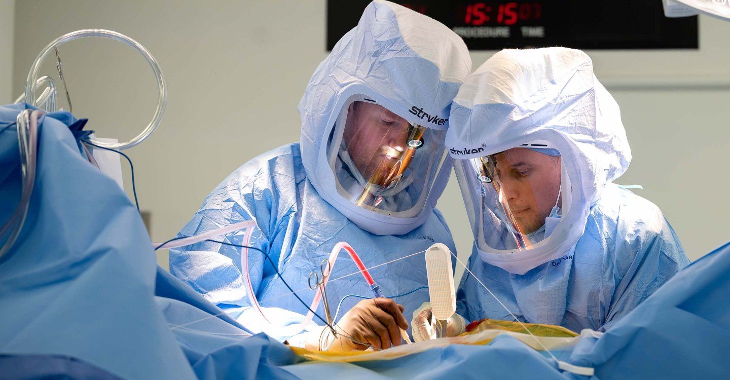 Two surgeons wearing overall protective gear operate on a patient. 