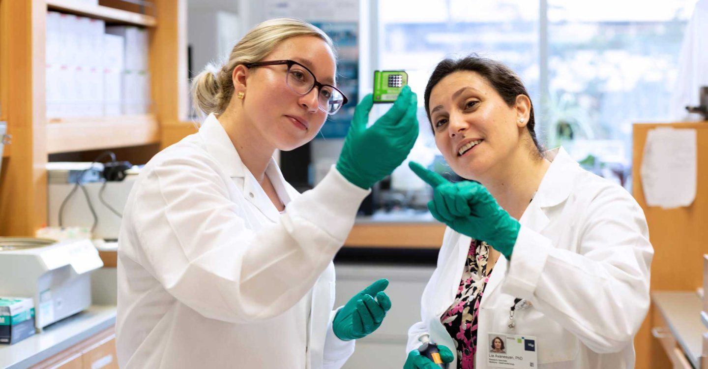 Two female researchers wearing lab coats and protective gloves examine a sample in a lab.