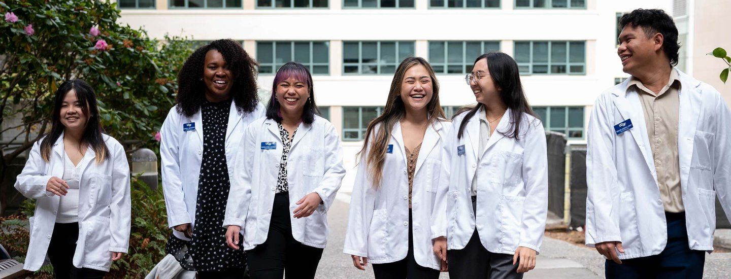 Students wearing white coats laugh with one another as they walk along a campus path