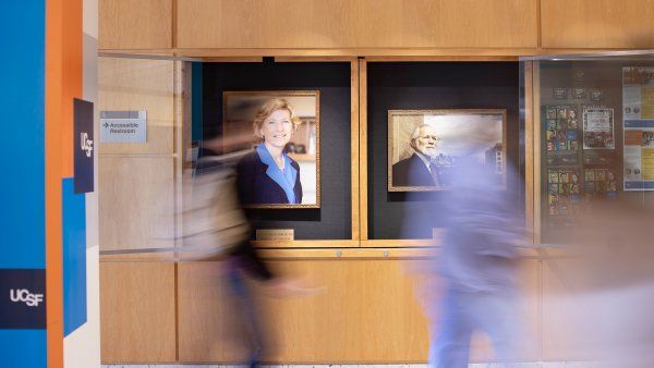people walk by the portraits of Susan Desmond-Hellmann and Michael Bishop in the hallway of the Medical Sciences Building on Parnassus Heights campus