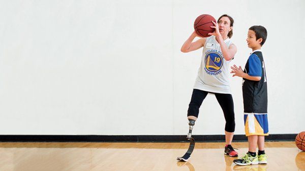Debrah Bevilacqua (left), has a prosthetic leg from the knee down and wears a Golden State Warriors shirt, shows a young child (right) how to play basketball indoors.