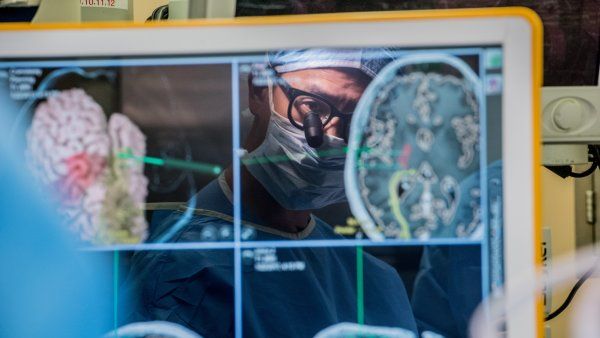 Neurosurgeon Edward Chang, dressed for surgery, looks through a semi-transparent monitor displaying brain activity.