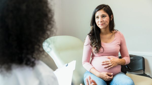 pregnant woman touches her abdomen as she listens to the unrecognizable female doctor. The doctor gestures as she holds the patient's medical records.
