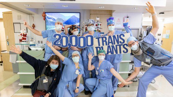 A group of medical professionals in medical scrubs holding a sign reading “20,000 transplants” pose happily for a group photo in a hospital hallway.