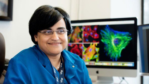 Shaeri Mukerjee wears a blue UCSF lanyard and a blue lab jacket while sitting in her office. Behind her is a monitor with microscopic images