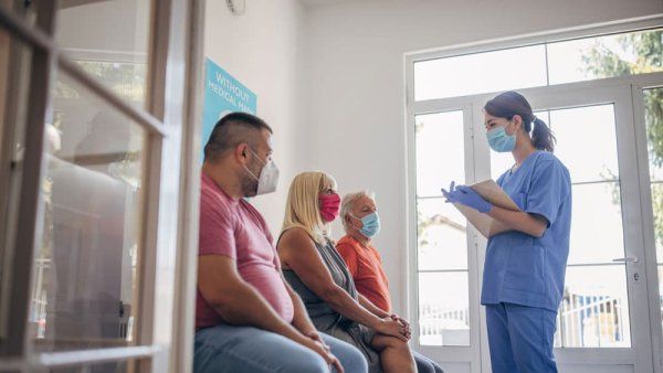 Three patients wearing masks listen to a medical professional in blue scrubs holding a clipboard