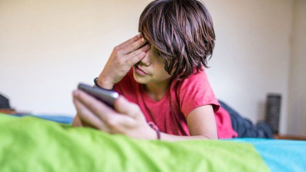 A pre-teen boy rubs his eye as he lays on his stomach while looking at his phone