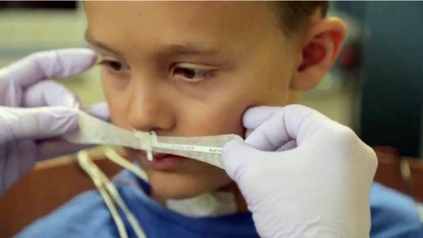 A pair of gloved hands place a nasal air sensor strip on the upper lip of a young boy.