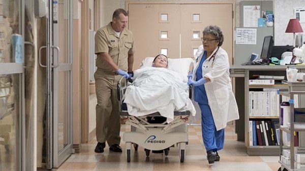 A uniformed officer and a doctor wheel a patient in a gurney down a hospital hallway