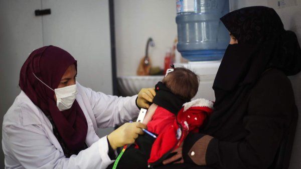 A doctor wearing a surgical mask examines a malnourished baby as her mother holds her.