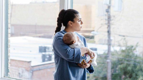 A mother holds her baby and looks contemplatively out of a large, light-filled window