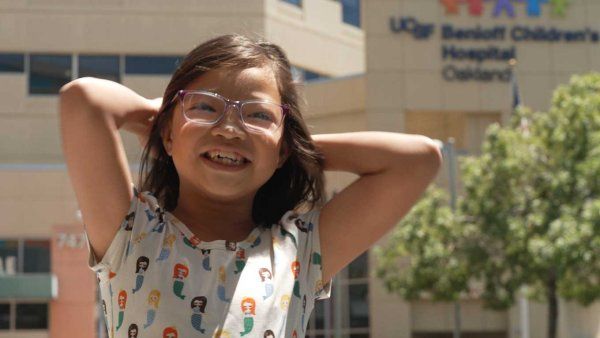 A small girl of Chinese descent smiles and places her arms behind her head as she stands in front of UCSF Benioff Children's Hospital, Oakland.