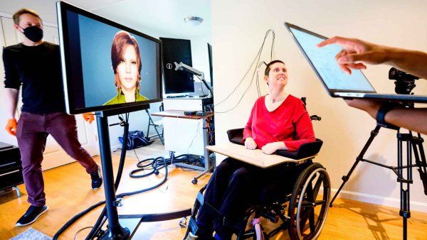 A woman wearing a red shirt and with a buzz haircut sits in her wheelchair and smiles. Next to her is a computer screen with a digital avatar that resembles her, and a researcher walks by.