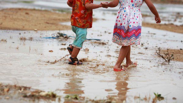 A toddler-aged boy and girl walk together through muddy water after strong rains.