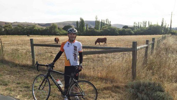 A middle-aged man stands with his bike in front of a pasture where a cow is grazing.