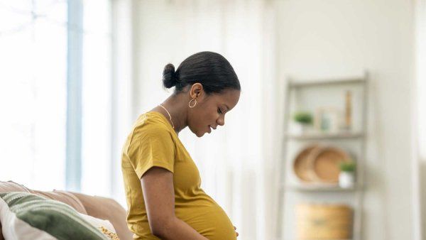 A Black pregnant woman sits comfortably on her couch at home and affectionately looks down at her growing stomach.