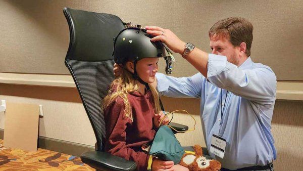 Kevin Bender (right) kneels as he adjusts a helmet on a young girl's head. She is sitting on an office chair, and the helmet will measure her eye movements.