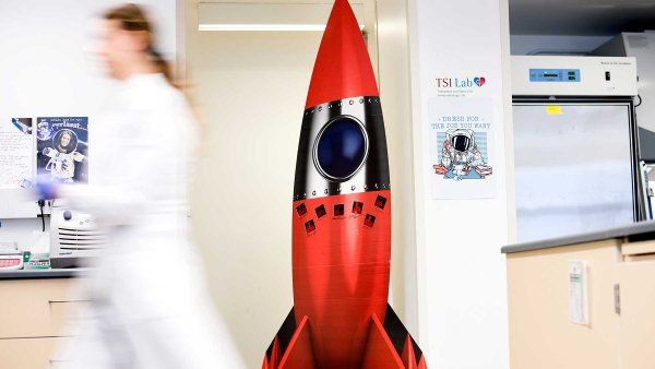 A large cutout of a red space rocket stands in the UCSF lab of Sonja Schrepfer and Tobias Deuse. A lab assistant in a white lab coat walks by.