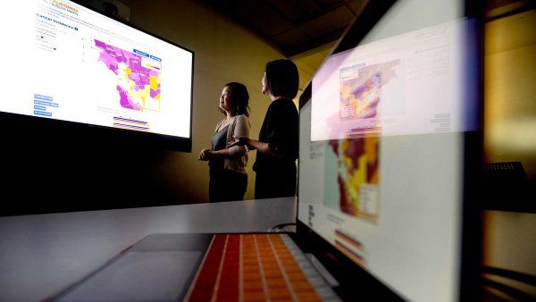 Scarlett Gomez and Debby Oh examine a screen with a map of the California Bay Area that displays cancer data. The room is dark, and in the foreground is a laptop, of which the screen reflects the researchers and the projection screen.