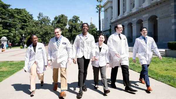 A group of medicine, pharmacy, and dental students wearing white coats smile as they walk together on a sunny pavement outisde the California State Capitol building in Sacramento.