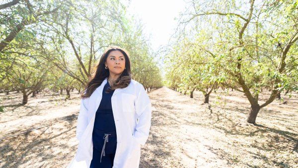Pharmacy technician Bibiana Arroyo wears a white coat as she walks through an agricultural orchard of trees in Modesto.